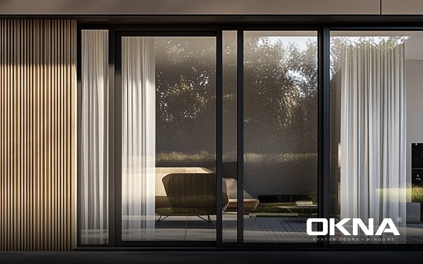 Modern sliding glass doors with semi-transparent curtains, indoor view to a garden at dusk.